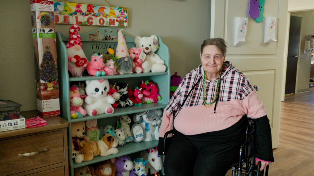 A woman sitting next to a shelf filled with her stuffed animals at a long term care facility