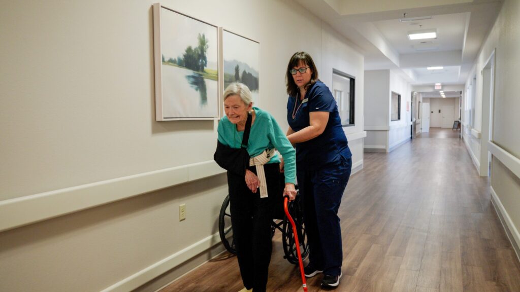 A nurse helps a woman down a hallway in a skilled nursing nursing home